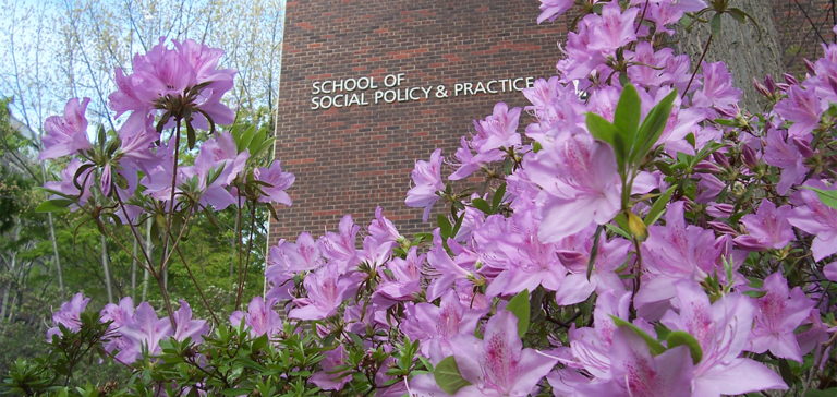 School of Social Policy & Practice sign on the Caster Building seen with pink azaleas.
