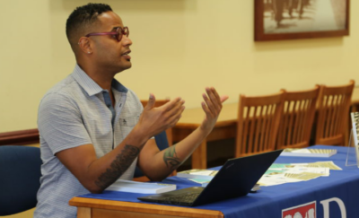 Ezekiel Dixon-Roman sits at a table with a laptop and speaks to an audience