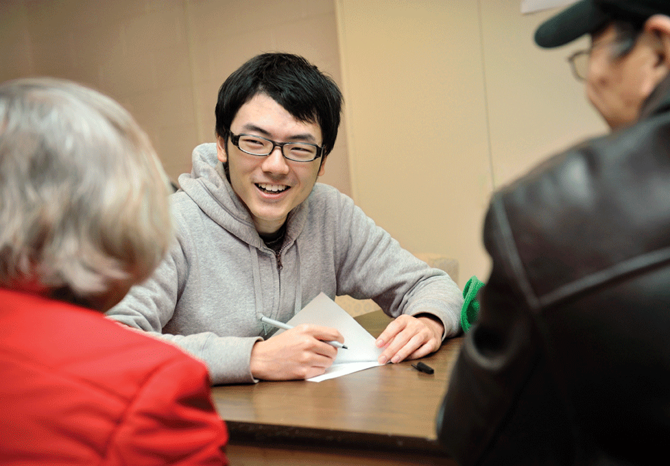 A man sitting at a desk holding a pen and paper, and smiling at two people who are out of focus