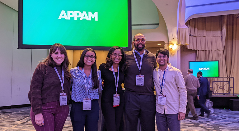 Five students and staff from SP2's MSSP program stand together smiling in a large room. A screen behind them says "APPAM."