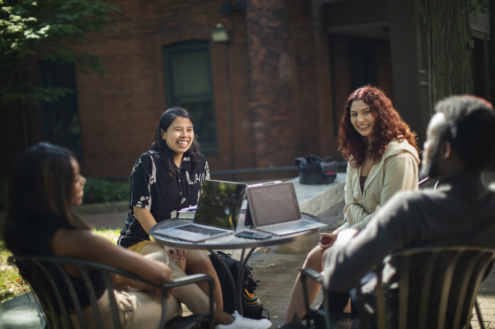 Four students sit smiling and conversing at a round table outdoors on campus. The photo is behind a transparent white layer on which text appears.