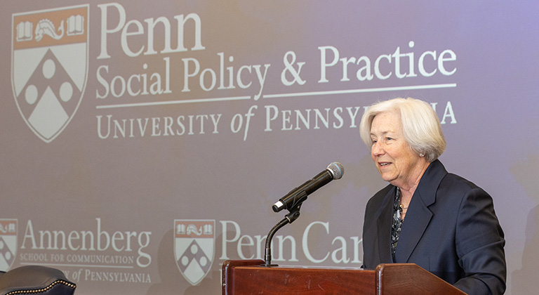 Dean Sally Bachman speaks at a podium in front of a blue screen that shows the logos of Penn's School of Social Policy & Practice, the Annenberg School, and Penn Carey Law.
