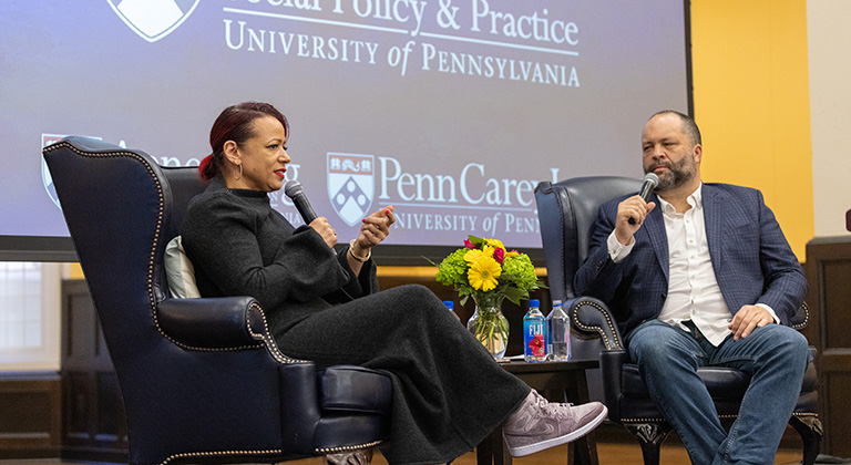 Nikole Hannah-Jones speaks and Ben Jealous listens as they sit onstage. A blue screen behind them shows the logos of the three sponsoring schools.