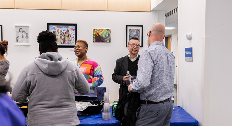 SP2 community members interact around a refreshment table near four framed works of art on a lobby wall.