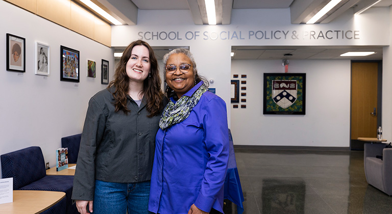 Rain Gideon and Dr. Jerri Bourjolly stand together smiling in the empty lobby. A row of framed artworks and "School of Social Policy & Practice" appear on the walls behind them.