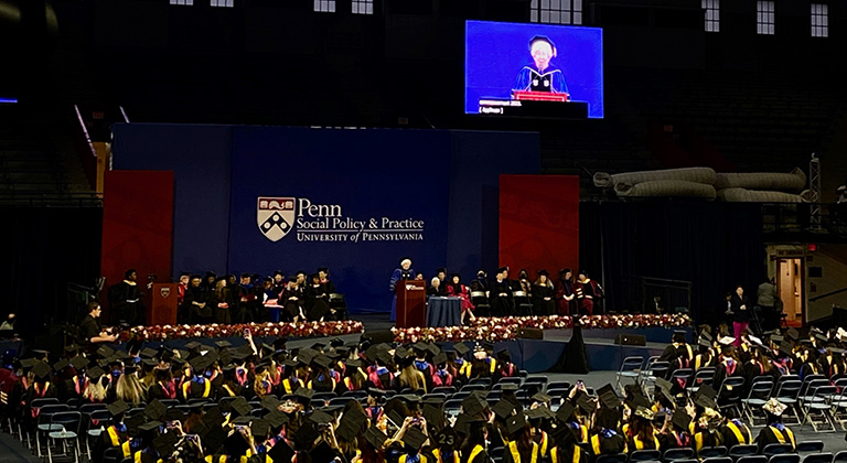 Dean Bachman speaks at a podium and projected onscreen before an audience of graduates at the Palestra, under the Penn Social Policy & Practice logo