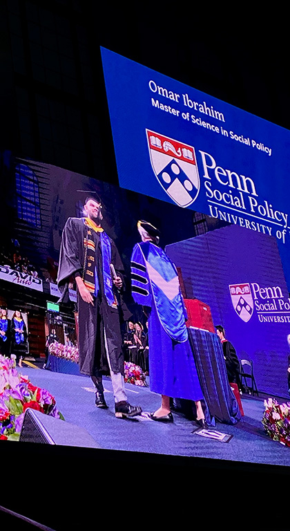 Two large screens show a graduate shaking the dean's hand onstage and the logo of Penn's School of Social Policy & Practice